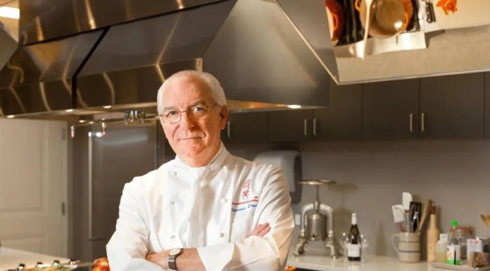 L’ Académie de Cuisine: Washington’s Gastronomic GEM An older male chef with glasses and short white hair stands confidently in a modern Washington kitchen. He wears a white chef’s coat, hinting at his gastronomic expertise, and smiles slightly amid sleek cabinets and stainless steel appliances.