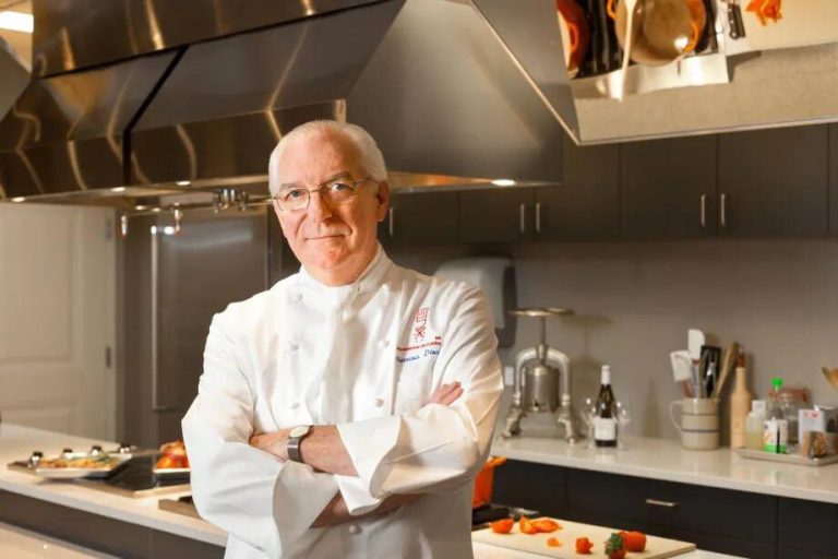Un chef cuisinier d'un certain âge, portant des lunettes et des cheveux blancs courts, se tient avec assurance dans une cuisine moderne de Washington. Il porte une blouse blanche de chef, ce qui laisse deviner son expertise gastronomique, et sourit légèrement au milieu des armoires élégantes et des appareils en acier inoxydable. 