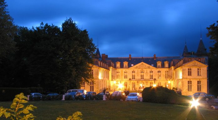 Les Hôtels Particuliers A grand, well-lit chateau evokes the charm of Hôtels Particuliers, with large windows and elegant architecture. Cars are parked in front, while luxury accommodation is framed by yellow-green bushes and trees beneath the twilight sky.