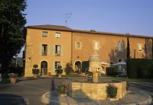 The Perignane Bastide A rustic Perignane bastide with ochre walls, blue shutters, and arched doorways stands behind a round stone fountain. Potted plants, topiary trees, white patio umbrellas, and chairs enhance the courtyard under a clear sky.