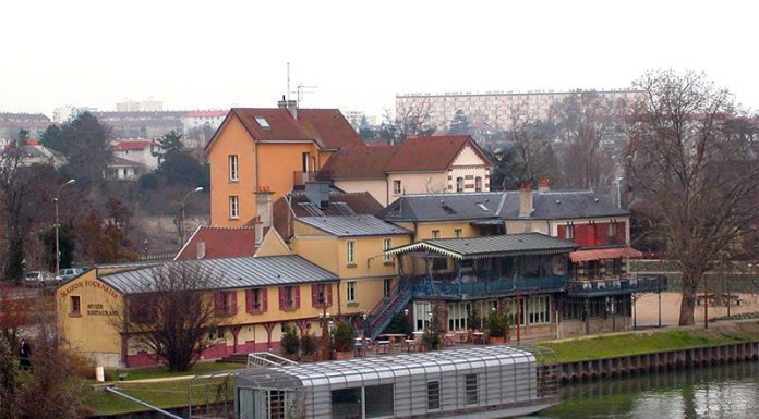 LES GUINGUETTES ET LA SEINE: A CELEBRATION OF RENOIR’S LUNCHON OF THE BOATING PARTY A metal-roofed houseboat is moored on a calm river, reminiscent of Renoir’s Seine scenes. Colorful buildings, including a yellow and red restaurant with outdoor seating, line the shore beneath a cloudy late autumn sky. Apartment blocks rise in the distance.