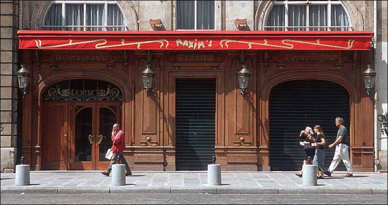 Vue de la rue du restaurant TWIN MAXIM'S, avec ses portes en bois ornées et ses volets noirs fermés. Un auvent rouge avec des accents jaunes et l'inscription MAXIMS couvre l'entrée, alors que quatre personnes passent devant, un homme à gauche, trois à droite. 