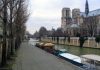 SOUNDS OF PARIS IN THE HEART OF THE VILLAGE A cobblestone path runs beside the Seine River in Paris, with trees lining the walkway. Several moored boats with yellow awnings sit on the river. Notre-Dame Cathedral, with its towers and gothic details, stands prominently on the right under a cloudy sky.