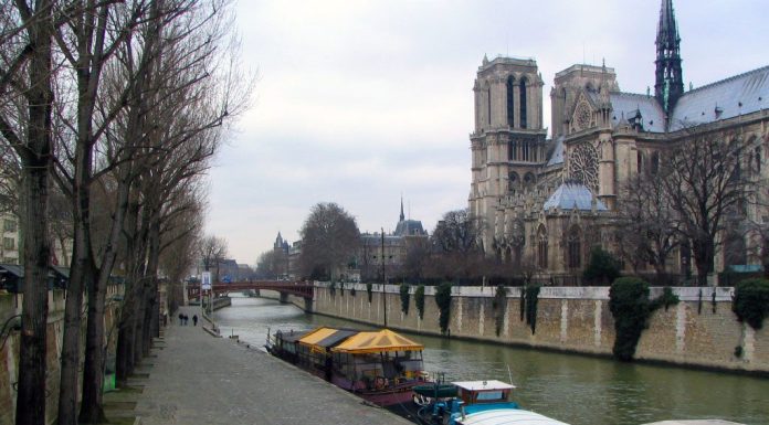 SOUNDS OF PARIS IN THE HEART OF THE VILLAGE View of the Seine River in Paris with houseboats moored along the banks, bare trees lining a cobblestone walkway, and the Gothic architecture of Notre-Dame Cathedral on the right—all set amid the charming sounds of Paris under a cloudy sky.