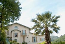 La Roseraie à Vence A beige two-story house with green trim and wooden shutters is surrounded by lush greenery in Vence. A tall palm tree stands in the foreground beneath the clear blue sky, while distant hills frame the charming La Roseraie residence.
