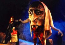 « La Couronne du Destin »: des marionnettes ou de vrais géants ? A person in a cloak stands beside a performer wearing a large, exaggerated troll marionnette with fangs and a big nose. The stage, set with rocky props and blue lighting, evokes the mystical world of La Couronne du Destin and its legendary géants.
