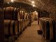 Why Does France Make the Best Wine? A dimly lit stone wine cellar with arched ceilings, lined on both sides with rows of large wooden barrels aging the best French wine. Warm lights illuminate rustic brick walls, creating a cozy atmosphere along the narrow path through the center.