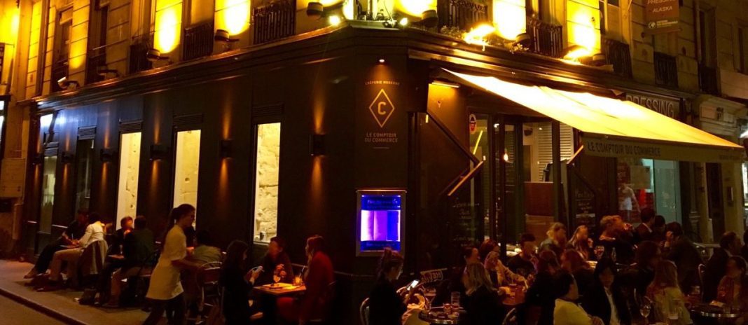le-comptoir-du-commerce A lively Parisian street café at night, Le Comptoir Du Commerce buzzes with diners at outdoor tables. Warm yellow lights illuminate the facade as a waiter serves guests and a glowing aquarium enhances the vibrant, bustling atmosphere near the entrance.