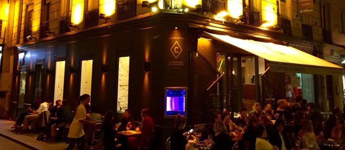 Café de rue parisien animé la nuit, Le Comptoir du Commerce est animé par des convives attablés à l'extérieur. Des lumières jaunes chaudes illuminent la façade pendant qu'un serveur sert les clients et un aquarium lumineux renforce l'atmosphère vibrante et animée près de l'entrée. 