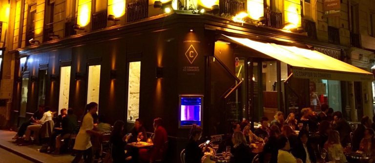 Café de rue parisien animé la nuit, Le Comptoir du Commerce est animé par des convives attablés à l'extérieur. Des lumières jaunes chaudes illuminent la façade pendant qu'un serveur sert les clients et un aquarium lumineux renforce l'atmosphère vibrante et animée près de l'entrée. 