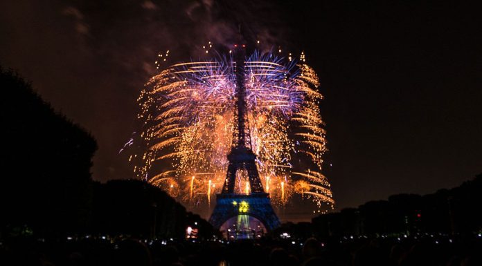 July 4th – July 14th Nighttime view of the Eiffel Tower in Paris, illuminated with vibrant fireworks bursting behind it on July 14th. The sky glows with gold and blue sparks as silhouetted crowds watch from the foreground, framed by trees on both sides.