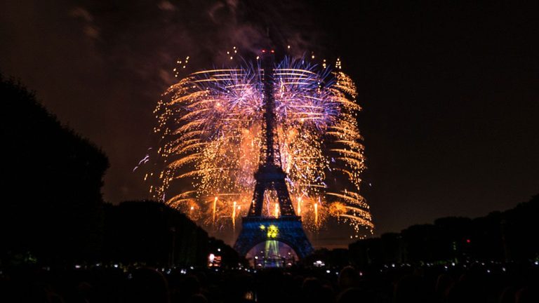 Vue nocturne de la Tour Eiffel à Paris, illuminée par un feu d'artifice éclatant derrière elle le 14 juillet. Le ciel s'illumine d'étincelles or et bleues, tandis que des silhouettes de foules observent le spectacle depuis le premier plan, encadrées par des arbres de part et d'autre. 