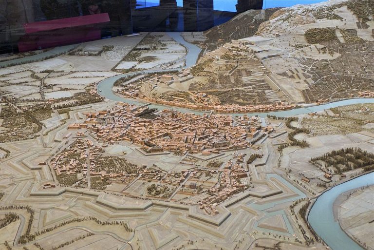 Une maquette détaillée dans le style des Plans-Reliefs de Vauban représente une ville historique entourée de murs défensifs et de bastions, de rivières sinueuses et de ponts ; des bâtiments compacts remplissent son centre, avec des collines, des routes, des champs et des arbres à l'extérieur.