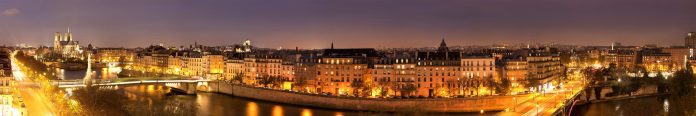 Vue panoramique nocturne de Paris avec des bâtiments illuminés bordant la Seine. La cathédrale Notre-Dame et l'île de la Cité sont visibles sur la gauche, tandis que les lampadaires dorés se reflètent sur l'eau sous un ciel clair et crépusculaire. 