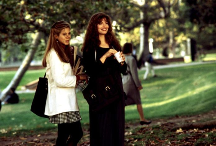 Deux femmes se tiennent dans un parc. L'une a de longs cheveux blonds, porte une veste blanche et une jupe à carreaux, et tient des livres étiquetés 