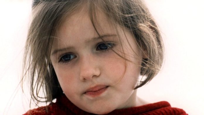 Gros plan d'une jeune fille à la peau claire et aux cheveux bruns raides, dont quelques mèches tombent sur son visage. Elle porte un pull-over rouge et regarde légèrement vers le bas avec une expression pensive et un peu triste. L'arrière-plan est légèrement flou et lumineux.  