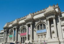 MUSEUMS A neoclassical museum building with large arched windows, tall Corinthian columns, decorative stonework, and banners hanging above the entrances. People are gathered on the wide steps in front, and the sky is clear and blue.