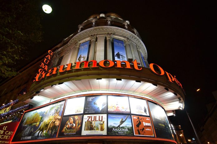 Vue nocturne du cinéma Gaumont Opéra à Paris, avec des enseignes au néon rouge vif et de grandes affiches de films placées au-dessus et au-dessous de l'entrée. La façade ornée du bâtiment est illuminée et la lune est visible dans le ciel sombre. 