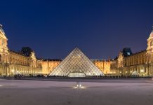 Le Louvre Night view of the Louvre Museum in Paris, featuring the illuminated glass pyramid entrance at the center, flanked by the historic museum buildings and a statue on the right side. The sky is dark blue, and the courtyard is mostly empty.