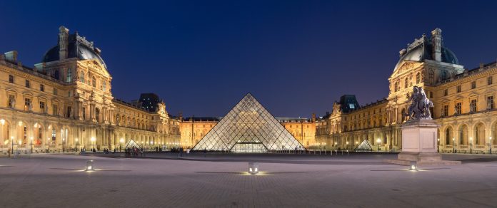 Vue nocturne du musée du Louvre à Paris, avec au centre l'entrée pyramidale en verre illuminée, flanquée des bâtiments historiques du musée et d'une statue sur le côté droit. Le ciel est bleu foncé et la cour est pratiquement vide. 