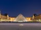 Le Louvre Night view of the Louvre Museum in Paris, featuring the illuminated glass pyramid entrance at the center, flanked by the historic museum buildings and a statue on the right side. The sky is dark blue, and the courtyard is mostly empty.
