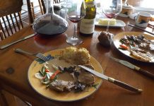 Traditional French Foods A wooden table set for a meal features a plate with meat in gravy, vegetables, and hash brown—reminiscent of Traditional French Foods. Wine glasses, a wine decanter, a bread roll, and butter complete the inviting scene.