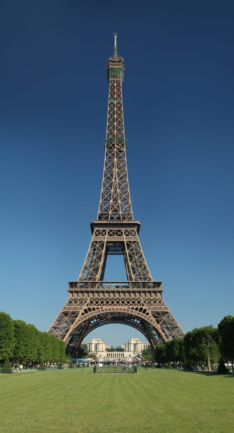 La tour Eiffel, emblème de Paris, se dresse dans un ciel bleu clair, entourée d'arbres verdoyants et d'une pelouse bien entretenue. Les gens se rassemblent près de la base, tandis que les bâtiments éloignés sont visibles à travers les pieds de la tour, ce qui ajoute à sa grandeur. 
