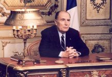 IN MEMORIAM : MY LIFE WITH MITTERAND An older man in a dark suit and tie sits at an ornate desk with a brass lamp, books, and papers. He clasps his hands thoughtfully, as if reflecting on my life; gold details and a French flag evoke the legacy of Mitterand in the lavish room.