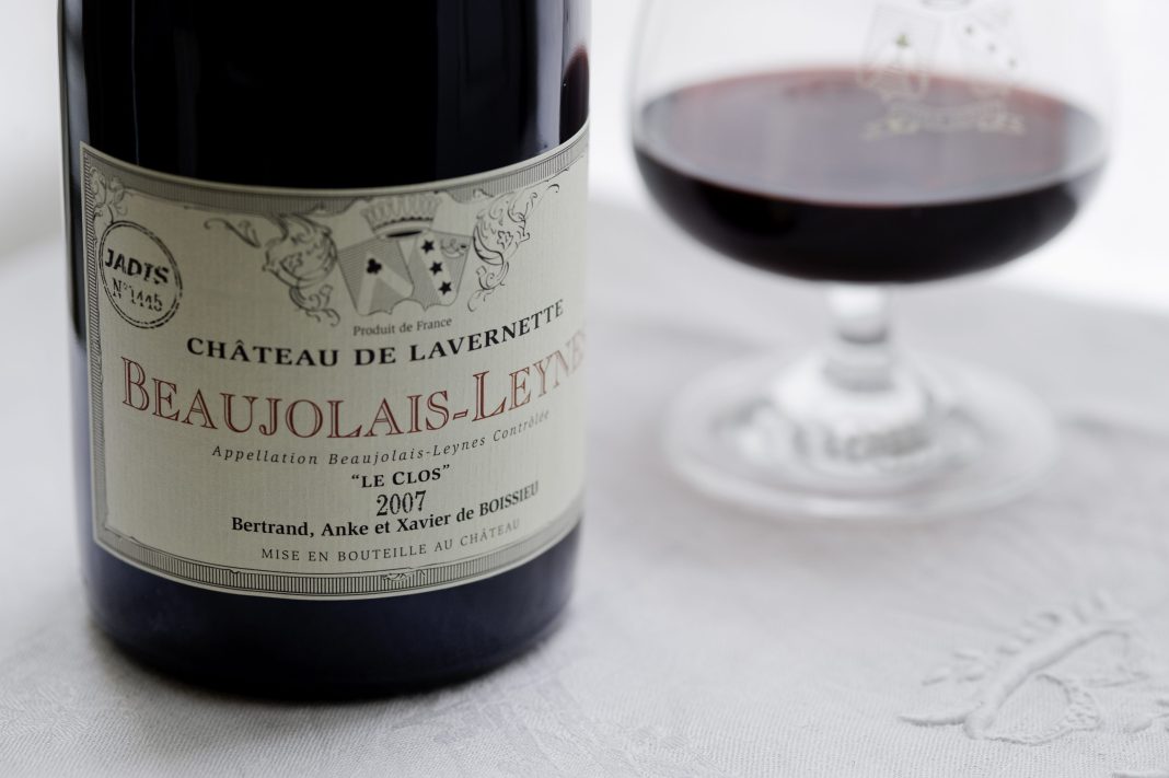 Gros plan sur une bouteille de vin étiquetée Château de Lavernette Beaujolais-Leynes 2007 à côté d'un verre de vin rouge sur une nappe blanche - une scène joyeuse célébrant le Beaujolais, avec des éléments décoratifs et les noms de Bertrand, Anke et Xavier de Boissieu.
