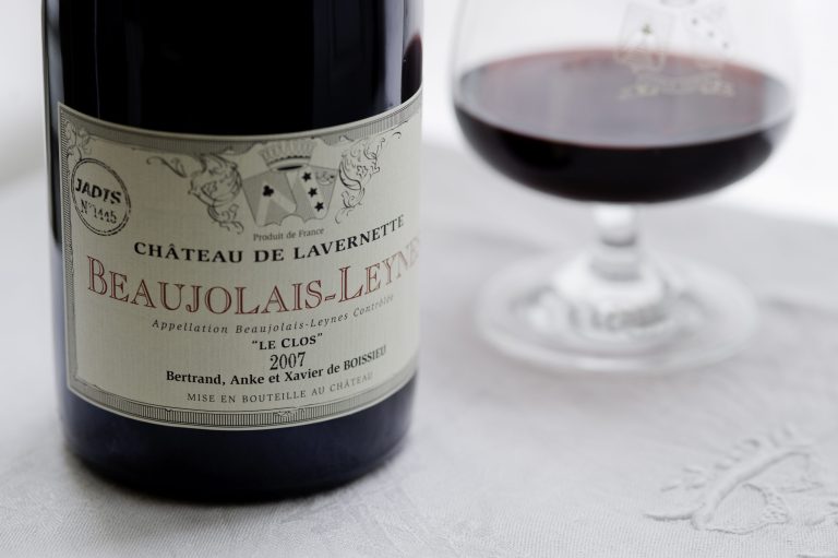 Gros plan sur une bouteille de vin étiquetée Château de Lavernette Beaujolais-Leynes 2007 à côté d'un verre de vin rouge sur une nappe blanche - une scène joyeuse célébrant le Beaujolais, avec des éléments décoratifs et les noms de Bertrand, Anke et Xavier de Boissieu.