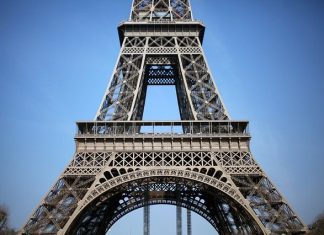 Le français: Une langue et un style de vie Close-up view of the lower section of the Eiffel Tower against a clear blue sky, showcasing its detailed iron lattice—a true icon of français style de vie. The photo captures the base and legs with intricate architectural patterns. No people visible.