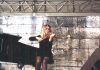 UN VIOLON, UN PIANO, DU JAZZ… A woman in a black dress sits on a stool, playing jazz on the violin on an outdoor stage. A grand piano is beside her, framed by large stone blocks in the background. Metal rigging and speakers surround the stage area.