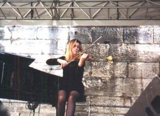 UN VIOLON, UN PIANO, DU JAZZ… A woman in a black dress sits on a stool, playing jazz on the violin on an outdoor stage. A grand piano is beside her, framed by large stone blocks in the background. Metal rigging and speakers surround the stage area.