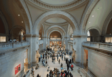 Dior, Proust et Diana A wide, high-ceilinged museum hall with elegant arches and stone walls is filled with many visitors walking, gathering, and observing. Natural light streams in from windows above, evoking a timeless elegance amid the busy, spacious interior.
