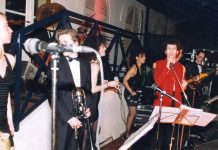 Elie de Rothschild Jr de toutes les fêtes A lively band performs onstage at the fêtes. A woman in a black dress stands at the left, a man in a tuxedo holding a trombone is near center, and another in a red jacket sings—evoking the elegance of an Elie de Rothschild event.