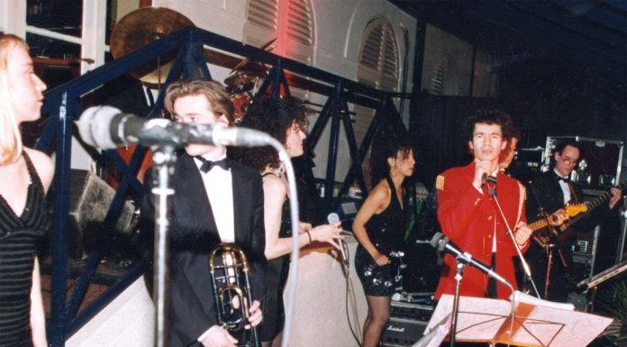 Elie de Rothschild Jr de toutes les fêtes A lively band performs onstage at the fêtes. A woman in a black dress stands at the left, a man in a tuxedo holding a trombone is near center, and another in a red jacket sings—evoking the elegance of an Elie de Rothschild event.