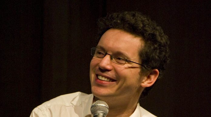 Les maths françaises encore à l’honneur A man with short curly dark hair and glasses smiles while holding a microphone. He is wearing a white button-up shirt, looking slightly to his left, ready to discuss maths françaises. The dark background puts full focus on him.