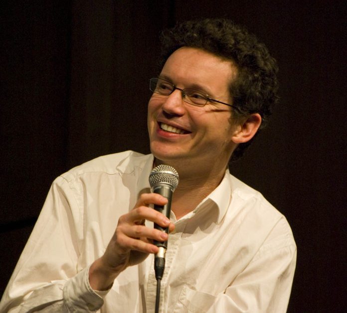 Un homme aux cheveux bruns courts et bouclés et aux lunettes sourit en tenant un microphone. Il porte une chemise blanche à boutons et regarde légèrement vers sa gauche, prêt à discuter de mathématiques françaises. L'arrière-plan sombre met l'accent sur lui.  