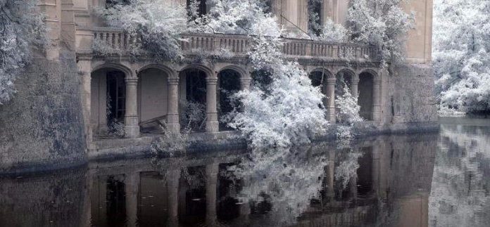 Ça vous dirait la VIE DE CHÂTEAU ? An old stone building with arched windows and a balcony stands partly submerged in calm water. Frost-covered trees surround this VIE DE CHÂTEAU scene, their icy foliage reflected below, creating an ethereal, wintry atmosphere.