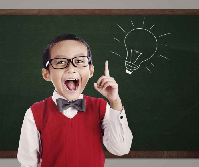 A gifted student wearing glasses, a red sweater vest, white shirt, and bow tie stands in front of a green chalkboard on Avenue de la Grande Armée. Smiling widely, he raises one finger beside a chalk drawing of a lit light bulb.