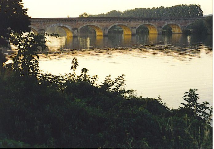 Un pont en arc de pierre enjambe une rivière calme au coucher du soleil en France, la lumière chaude se reflétant sur l'eau. Des arbres verdoyants encadrent la scène, ce qui en fait une vue idyllique souvent admirée lors d'une excursion en péniche de luxe dans cette campagne tranquille. 