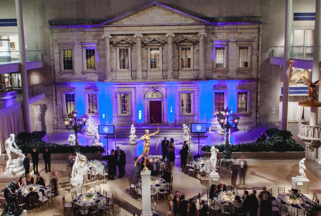 metropolitan-museum-of-art A grand indoor hall with an ornate, classical building facade lit by blue lights. Elegant round tables and chairs are set for an event. People in formal attire mingle among white marble statues and a golden statue at the center. The atmosphere is sophisticated and festive.