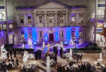 News A grand indoor hall with an ornate, classical building facade lit by blue lights. Elegant round tables and chairs are set for an event. People in formal attire mingle among white marble statues and a golden statue at the center. The atmosphere is sophisticated and festive.