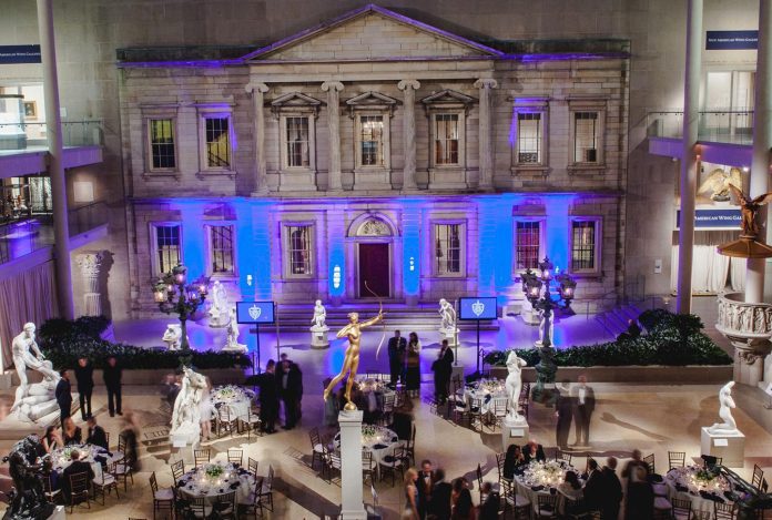 A grand indoor hall with an ornate, classical building facade lit by blue lights. Elegant round tables and chairs are set for an event. People in formal attire mingle among white marble statues and a golden statue at the center. The atmosphere is sophisticated and festive.