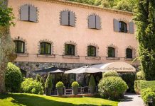 Hostellerie des Gorges de Pennafort A rustic, two-story stone building with gray shutters and arched windows houses the Hostellerie des Gorges de Pennafort hotel and restaurant. Lush greenery surrounds shaded outdoor seating with black umbrellas for guests to enjoy.