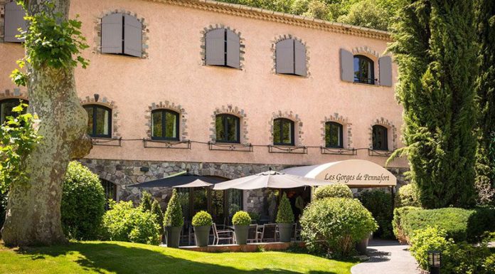 Hostellerie des Gorges de Pennafort A rustic, two-story stone building with gray shutters and arched windows houses the Hostellerie des Gorges de Pennafort hotel and restaurant. Lush greenery surrounds shaded outdoor seating with black umbrellas for guests to enjoy.