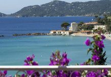 Hotel Belle-Vue A scenic coastal view with clear blue water, a sandy beach lined with sun loungers, and small white buildings by the shore—including Hotel Belle-Vue. Purple bougainvillea blooms in the foreground, with forested hills rising under a clear sky.