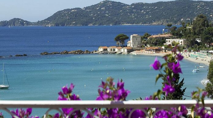 Hotel Belle-Vue A scenic coastal view with clear blue water, a sandy beach lined with sun loungers, and small white buildings by the shore—including Hotel Belle-Vue. Purple bougainvillea blooms in the foreground, with forested hills rising under a clear sky.