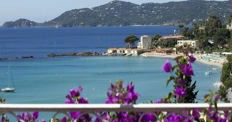 Vue pittoresque de la côte avec une eau bleue claire, une plage de sable bordée de chaises longues et de petits bâtiments blancs sur le rivage, dont l'hôtel Belle-Vue. Des bougainvilliers violets fleurissent au premier plan, tandis que des collines boisées s'élèvent sous un ciel clair. 
