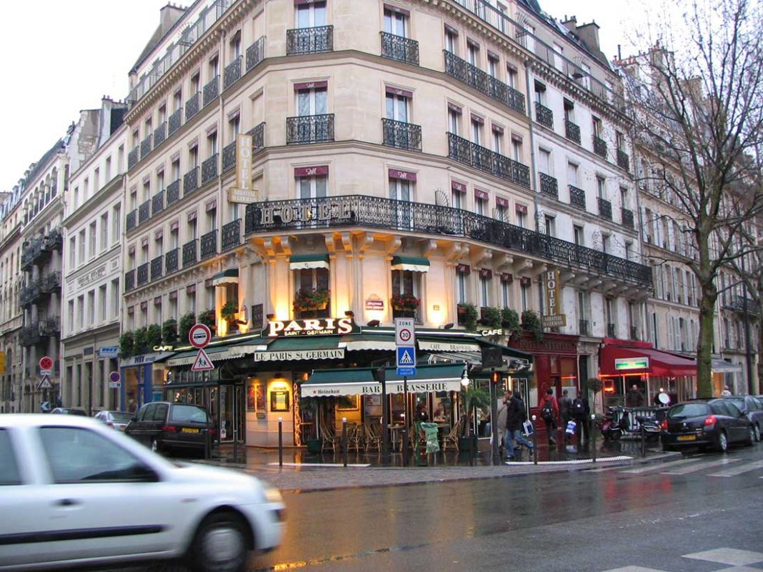 A bustling Paris street corner in the Latin Quarter features a classic cream-colored building with ornate balconies. At ground level, the lively café “Paris” boasts green awnings, outdoor seating, and people dining as cars and pedestrians navigate the wet street.